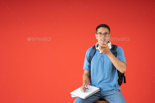 Male student with glasses, talking on speaker phone, holding a notebook and pencil