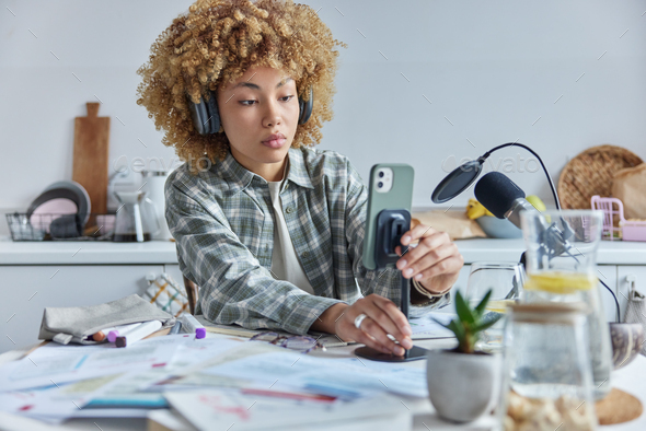 Serious curly haired female student studies online uses smartphone and microphone sits at table with - Stock Photo - Images