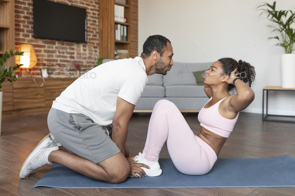 Happy young black husband helping to wife in sportswear doing abs exercises, kissing on mat - Stock Photo - Images