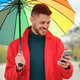 Latin young man with a red coat and a rainbow umbrella using a phone in autumn. - PhotoDune Item for Sale