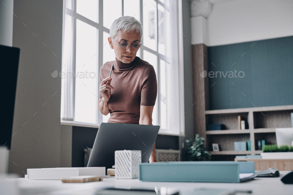 Senior businesswoman looking thoughtful while standing near her working place in office - Stock Photo - Images