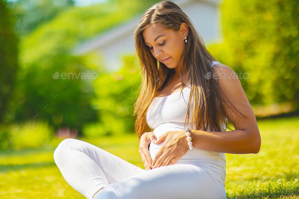 Beautiful pregnant woman sitting outdoor making hand heart gesture on belly - Stock Photo - Images