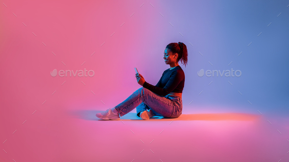 Amazing application. Happy black lady using smartphone, sitting on studio background in neon light