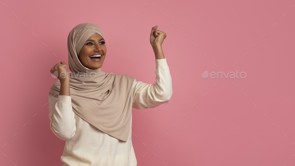 Overjoyed muslim woman celebrating success, shaking fists and looking aside