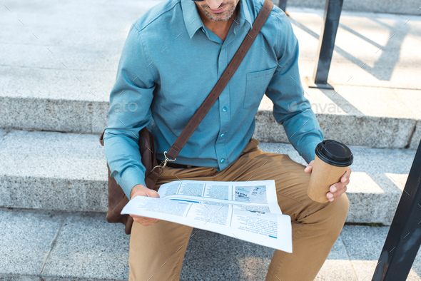 cropped shot of businessman with coffee to go sitting on stairs and reading newspaper