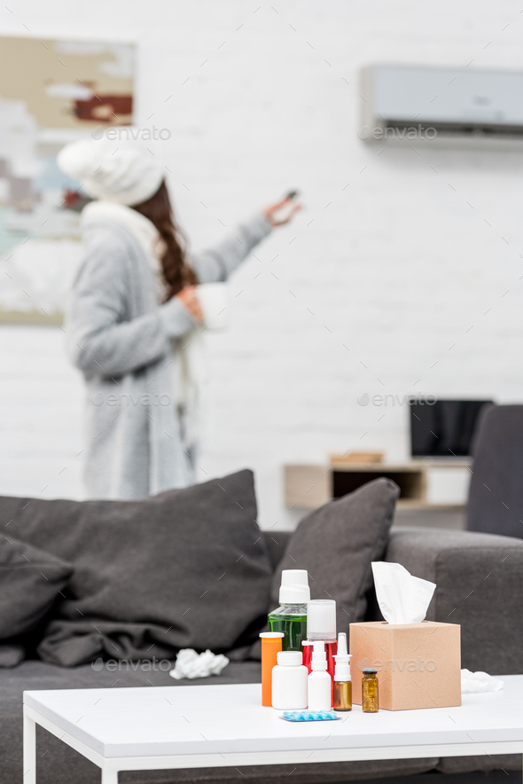 sick woman in warm clothes pointing at air conditioner with remote with various medicines standing
