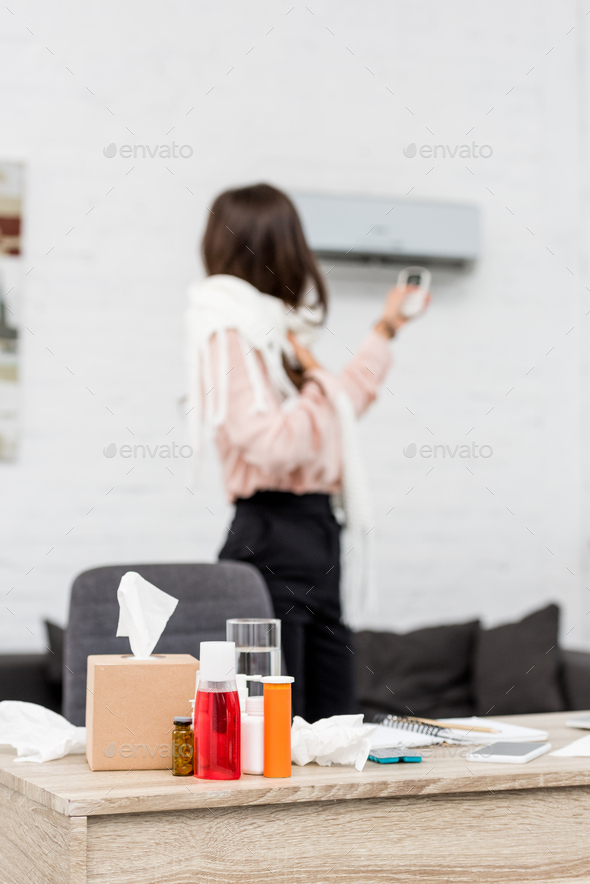 sick young woman pointing at air conditioner with remote control with medicines on foreground
