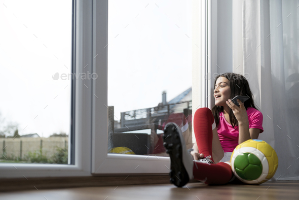 Girl in soccer outfit sitting on floor in living room speaking to someone on her smartphone