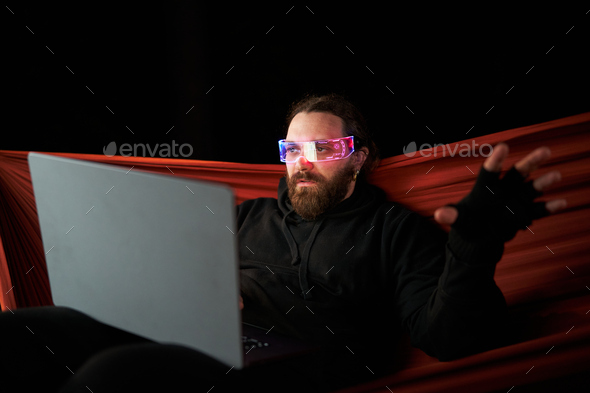 Gamer using a laptop and a extended Reality goggles while sitting on an hammock at night
