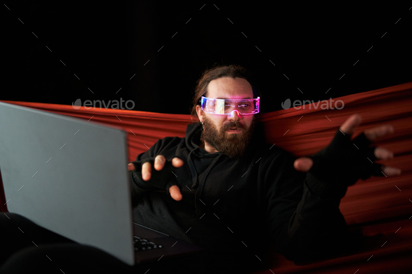 Man sitting on an hammock pointing while playing with a laptop and VR goggles