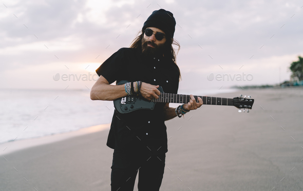 Long haired rock star in sunglasses using folk guitar for playing summer melody at seashore