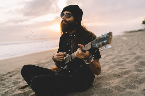 Carefree man in sunglasses using folk guitar for playing summer melody at seashore
