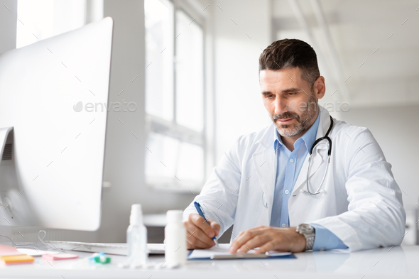 Serious male doctor using computer and writing notes in medical journal sitting at workplace in