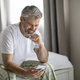 Cheery mature man sitting on bed, using smartphone and smiling - PhotoDune Item for Sale