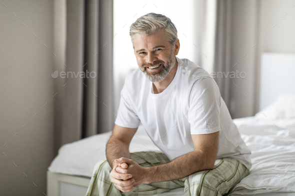 Portrait of happy middle aged man sitting on bed, smiling - Stock Photo - Images