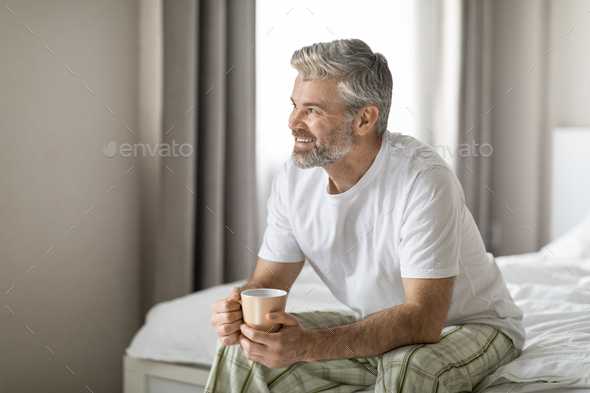 Joyful middle aged man sitting on bed, drinking coffee - Stock Photo - Images
