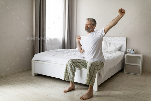 Happy handsome middle aged man sitting on bed and stretching - Stock Photo - Images