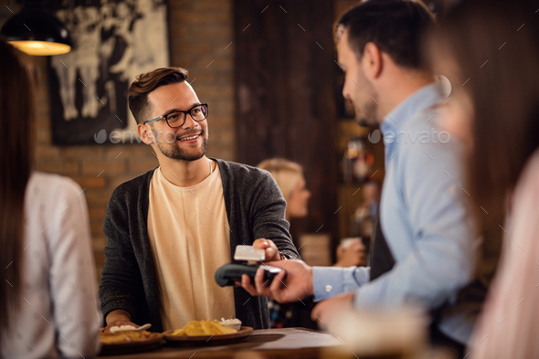 Young smiling man using mobile phone while paying a bill in a restaurant.