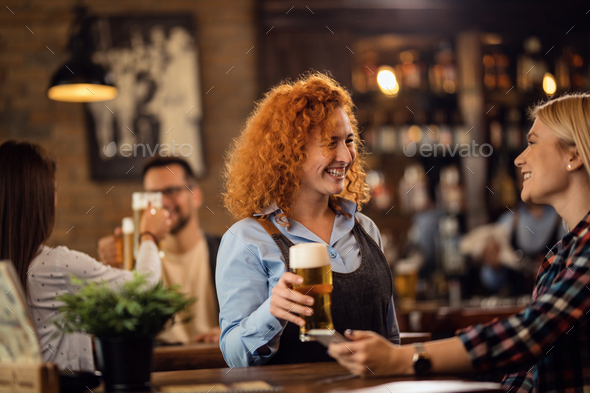 Happy waitress talking to a woman while giving her a glass of beer in a pub.