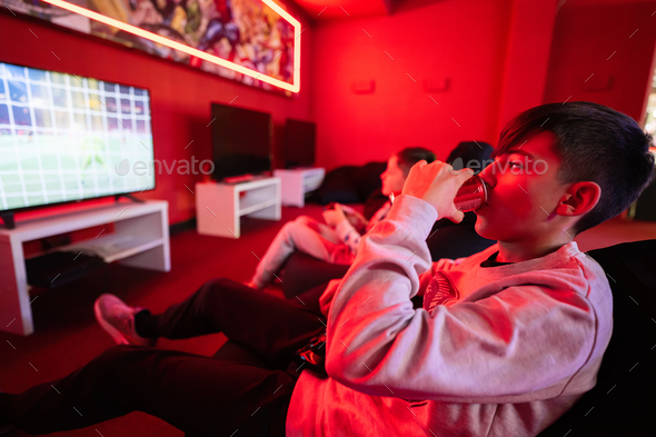 Two boys gamers play football gamepad video game console in red gaming room. Drink soda can.