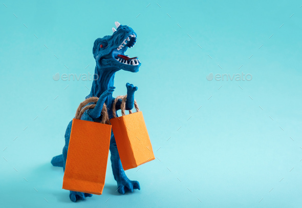 Cute blue dinosaur with orange shopping bags on a blue background.  - Stock Photo - Images