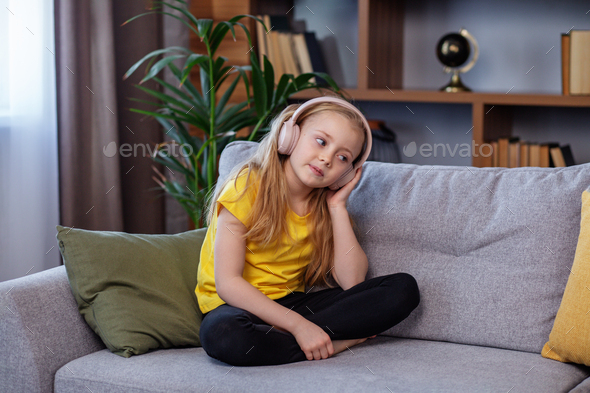 Happy child listens to music or audiobook at home on couch. Little school kid wearing headphones - Stock Photo - Images