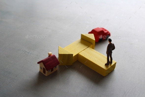 Miniature man standing at a turning point with car and house.