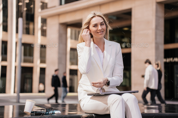 Cheerful young blonde entrepreneur woman sits with laptop with happy facial expression puts earphone