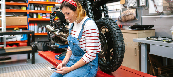 Mechanic woman looking phone sitting over platform with motorcycle on factory - Stock Photo - Images