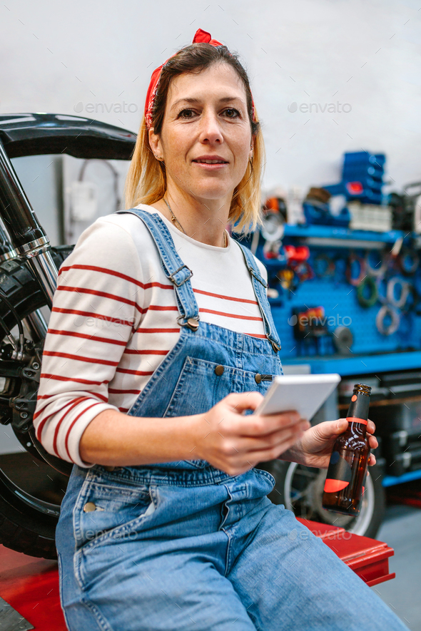 Mechanic woman looking phone and holding beer sitting in front of motorcycle on factory - Stock Photo - Images