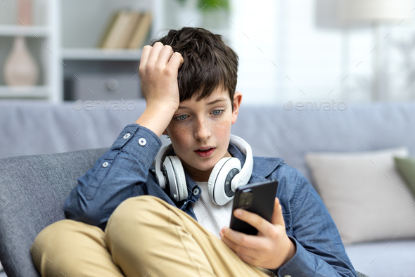 Young teenager boy reading bad news on phone, son sitting on sofa in living room with headphones at