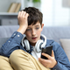 Young teenager boy reading bad news on phone, son sitting on sofa in living room with headphones at - PhotoDune Item for Sale