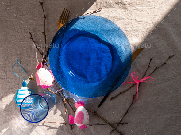 Happy Easter table setting blue glass plate fork knife tree branches on linen cloth. Festive menu