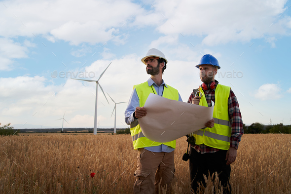 Two engineers in hard hats work holding plane in agricultural field with wind turbines. Outdoors.