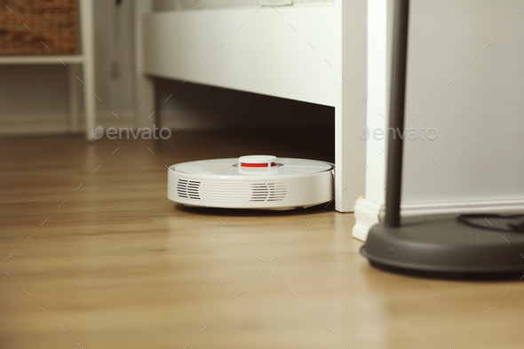 White robotic vacuum cleaner on laminate floor cleaning dust in living room interior. Smart electro