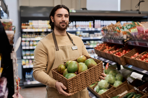 Young man working in supermarket and carrying box with fresh exotic fruits