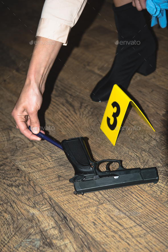 cropped view of hand examining gun near evidence marker at crime scene