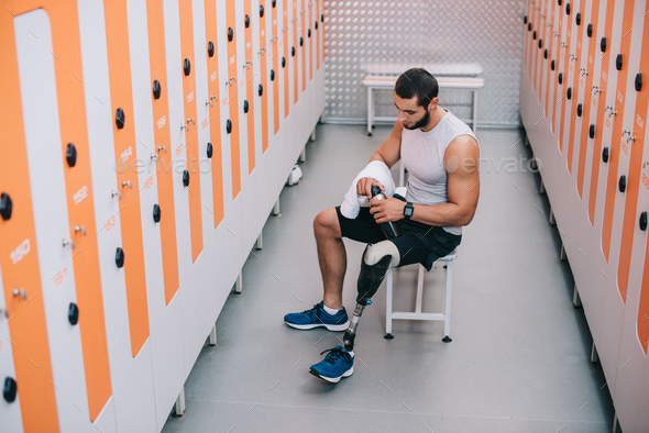 handsome young sportsman with artificial leg sitting on bench at gym changing room