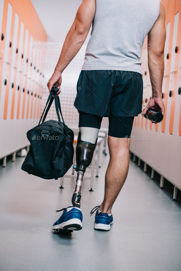 cropped shot of sportsman with artificial leg standing at gym changing room with bottle and bag