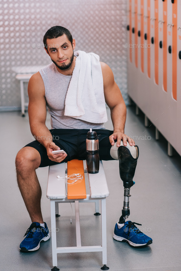 handsome young sportsman with artificial leg sitting on bench at gym changing room and using