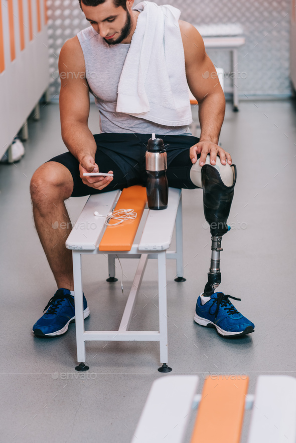 attractive young sportsman with artificial leg sitting on bench at gym changing room and using