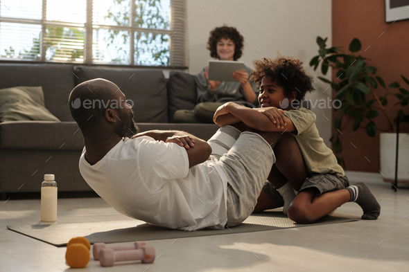 Cute little boy helping his father with sit ups while holding his