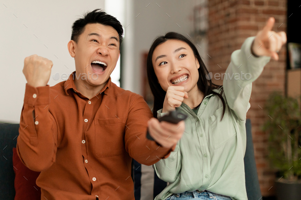 Joyful asian spouses shaking fists and shouting, watching sport channel, woman pointing on TV
