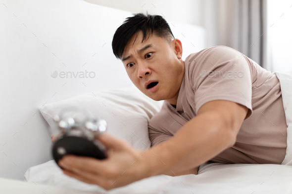Shocked overslept asian man lying in bed, looking at alarm