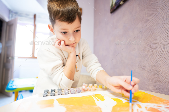 Left handed boy is painting by numbers with gouache at home.