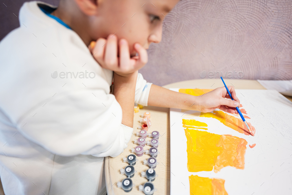 Left handed boy is painting by numbers with gouache at home.