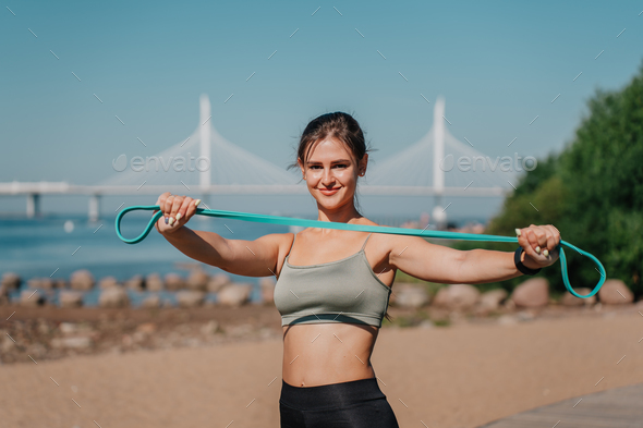 brunette hispanic girl in sportswear stretches rubber tapes, doing workout at beach with view on bay