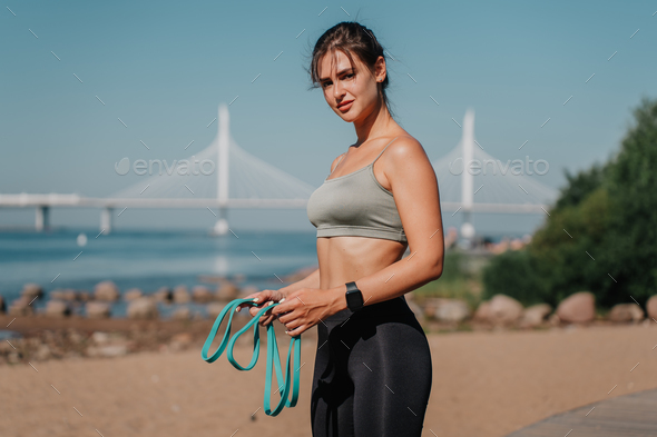 brunette hispanic girl in sportswear holds rubber tapes, doing workout at beach with view on bay