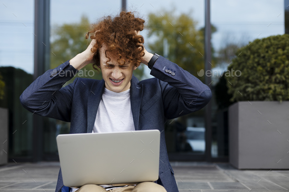 Worried guy holding his head, using laptop, sitting stairs near modern office.