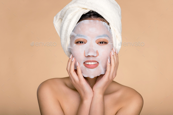 Cosmetic skin care procedure. Smiling pretty Asian young woman with facial cotton moisturizing mask